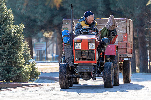 Bishkek, Kyrgyzstan - December 29, 2025: An elderly man in a blue hat and yellow vest sits in an orange mini tractor, which is stopped on a park pathway. He is looking at his smartphone, using modern technology in a park setting.  The photo captures a moment of rest or a break from work.