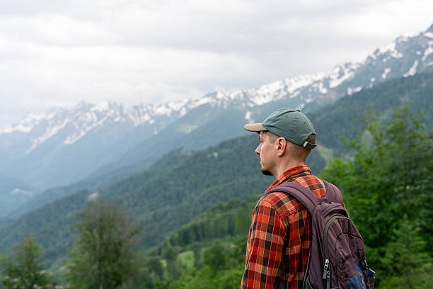 istock Side view of man with backpack standing on mountain viewpoint , looking at scenic alpine landscape with forested hills and snow-capped peaks in distance Concept of hiking, adventure travel copy space 2254063087