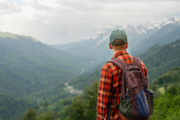male hiker wearing red shirt, carrying backpack and cap, enjoys panoramic mountain view during hike. Green valleys, forests, and snow-capped peaks, travel, exploration, active lifestyle, copy space