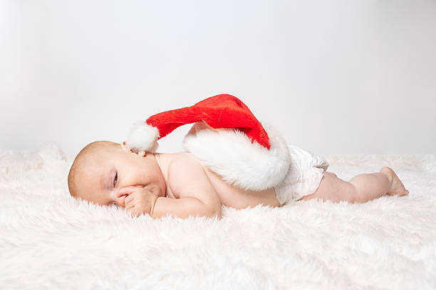 An adorable 6 month old Caucasian baby boy with a Santa hat on his back as he sucks his thumb and tries to stay awake with one eye peeking.