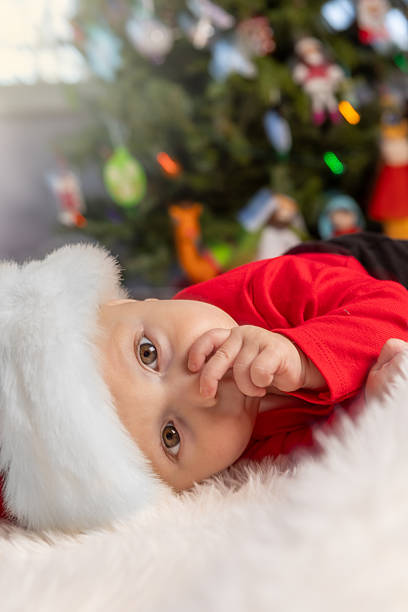 A cute 6 month old baby boy in a Santa hat sucking his thumb on the floor by the Christmas tree.