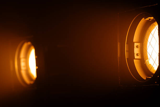Two powerful studio spotlights emit warm orange beams in the darkness, creating a striking cinematic lighting effect. Lighting equipment backstage.