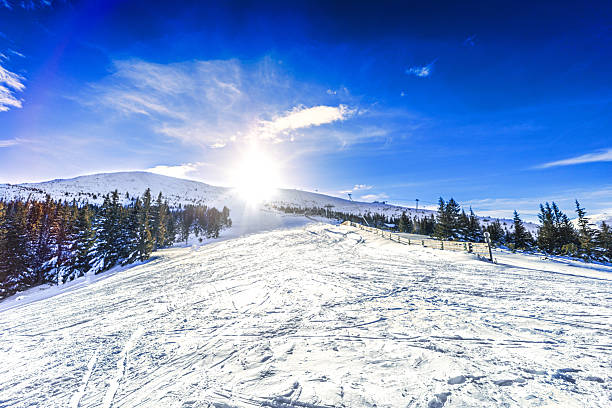 A wide scenic shot of a groomed ski slope in the Low Tatras, Slovakia, under a dramatic cloudy sky during twilight, featuring snow-covered ridges and evergreen forests.