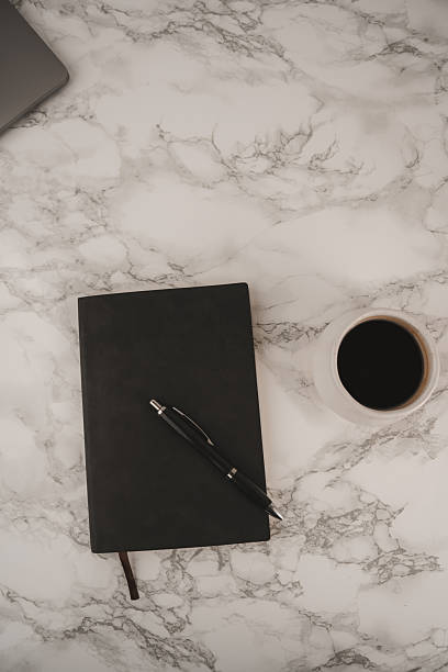 istock A black notebook lies open beside a sleek pen on a smooth marble surface. Next to it sits a white cup filled with dark coffee, inviting moments of inspiration and creativity 2254070118