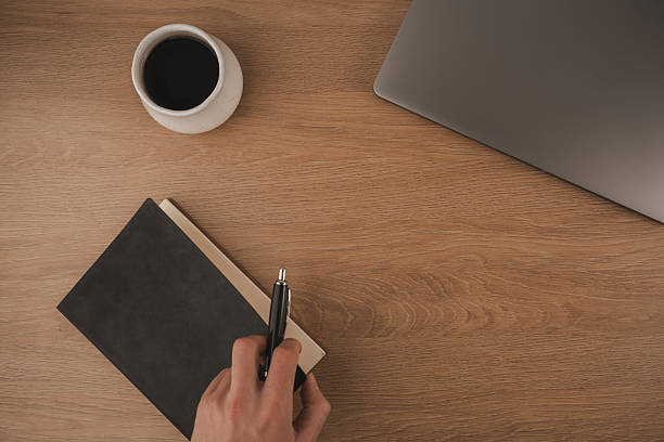 istock A hand writes notes in a notebook at a home workspace. A cup of coffee sits nearby, next to a laptop on a wooden surface, creating a focused atmosphere for productivity 2254070252