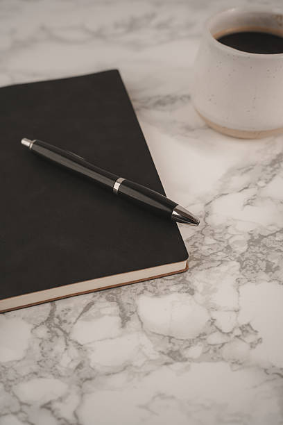 istock A black notebook lies open with a pen resting on top, accompanied by a white coffee cup filled with dark coffee. This setup invites creativity and reflection 2254070595