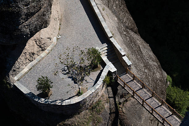A top-down perspective of a traditional stone-paved observation deck and a wooden bridge built into the cliffs of Meteora, Greece. The image showcases the unique religious architecture and dramatic rock formations under bright sunlight.