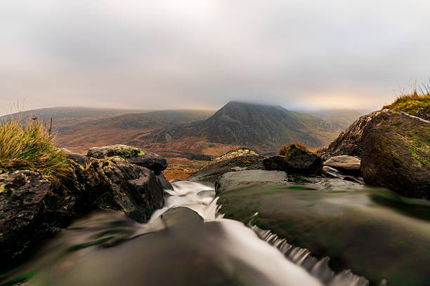 Water cascading over mossy rocks with a dramatic mountain landscape and cloudy sky in Snowdonia, Wales