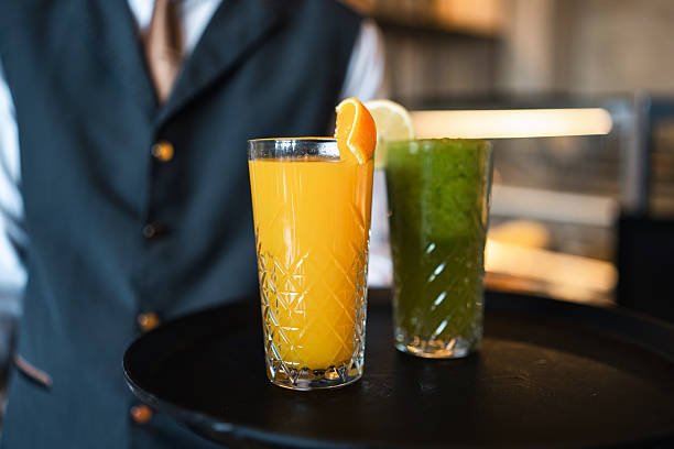 Waiter in formal attire carrying a tray with freshly squeezed orange and green juices garnished with citrus slices at a modern restaurant setting.