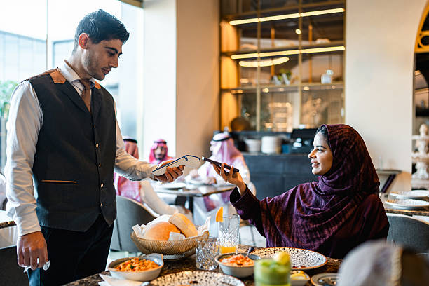 Young Arab woman in fine dining restaurant cheerfully using smartphone to make payment while interacting with waiter holding card reader.
