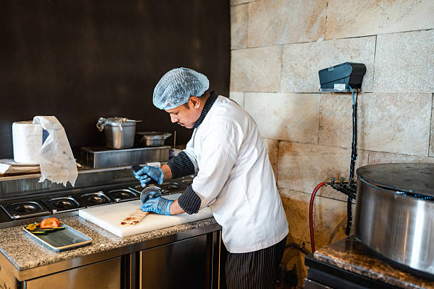 Mid adult Arab chef in professional kitchen slicing ingredients on cutting board with focused attention while wearing protective gear and surrounded by culinary tools.