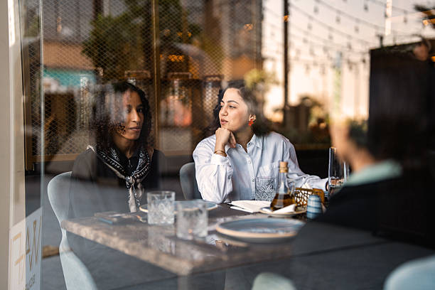 Two adult women sharing drinks and conversation inside a urban cafe, seen through the window with city reflections