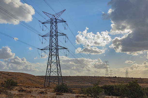 A high voltage electricity transmission tower stands tall in a rural landscape, supporting multiple overhead power lines that distribute electrical energy across long distances. The steel lattice structure is photographed from a low angle against a dramatic sky with clouds and blue tones, emphasizing scale, infrastructure strength, and energy distribution. The image represents power generation, national grid systems, electrical engineering, and modern energy infrastructure with clear copy space for design and editorial use