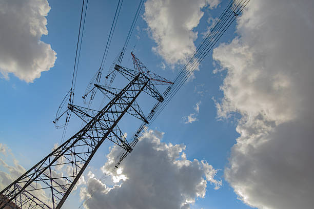 A high voltage electricity transmission tower stands tall in a rural landscape, supporting multiple overhead power lines that distribute electrical energy across long distances. The steel lattice structure is photographed from a low angle against a dramatic sky with clouds and blue tones, emphasizing scale, infrastructure strength, and energy distribution. The image represents power generation, national grid systems, electrical engineering, and modern energy infrastructure with clear copy space for design and editorial use