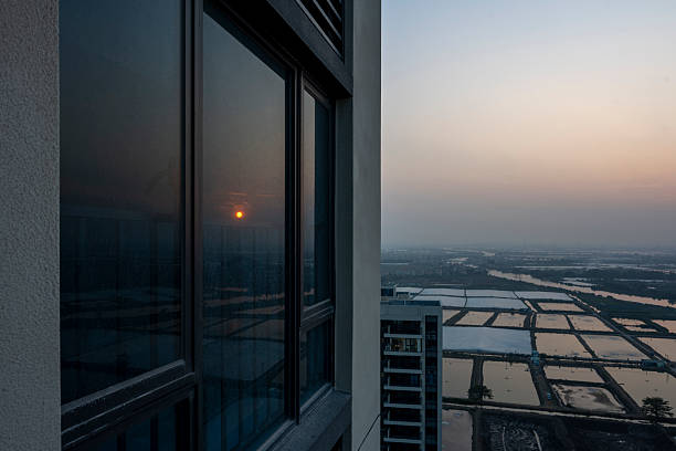 Captivating urban sunset view from a high-rise building overlooking agricultural fields. The image captures the serene blend of modern architecture and rural landscape during dusk, offering a tranquil yet dynamic visual experience suitable for real estate marketing or travel photography.