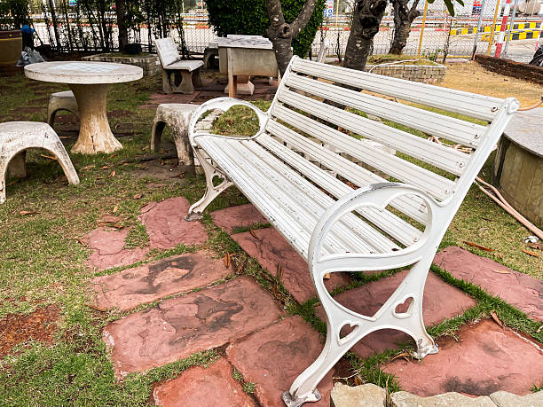 A white metal park bench placed on a stone pathway in a small garden area, surrounded by outdoor tables and chairs, creating a calm and relaxing public space.