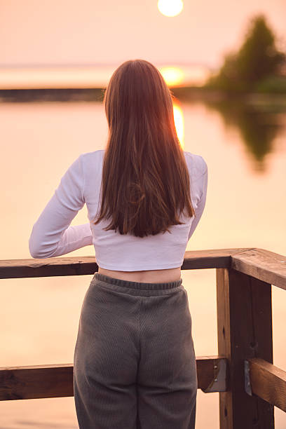 Young woman with long brown hair gazes at sunset over calm lake. Soft pink and orange hues fill sky, creating serene ambiance. Wooden deck provides peaceful view
