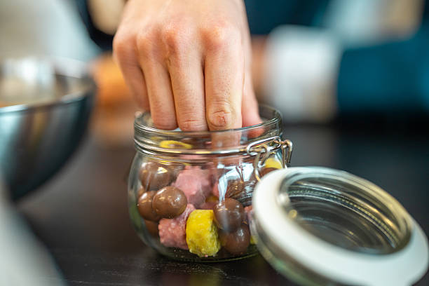 Businessman in office environment reaching hand into glass jar filled with colorful chocolate candies during a work break on desk.