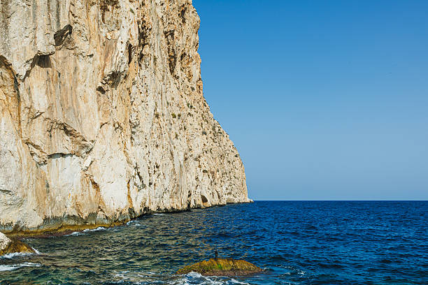 Majestic cliffs rise sharply from the sparkling blue water, with sunlight illuminating the rocky surfaces. The calm sea stretches endlessly beneath a bright, cloudless sky.