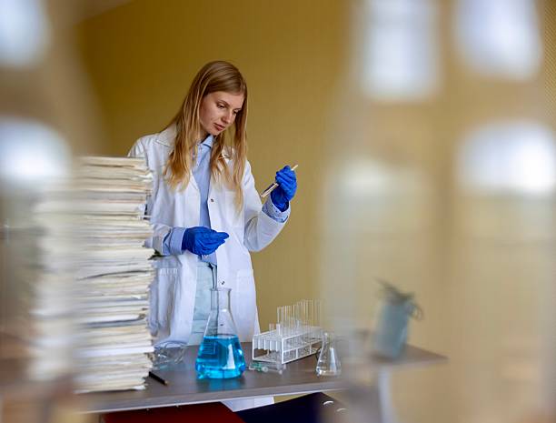 A young Caucasian woman with blonde hair, wearing a white lab coat and blue nitrile gloves, focuses intently on pipetting a blue liquid from a beaker into a test tube in a contemporary laboratory setting in Belgium. She stands at a lab bench cluttered with stacked papers, additional test tubes in a rack, and glassware, conveying a mood of concentration and scientific precision during an experiment. The indoor environment features soft yellow walls and blurred background elements, suggesting a professional research facility. This image is suitable for commercial uses such as illustrating scientific research articles, biotechnology advertisements, educational materials on laboratory techniques, medical journals, and corporate profiles in pharmaceuticals or chemistry, highlighting themes of innovation and hands-on science in a European context.