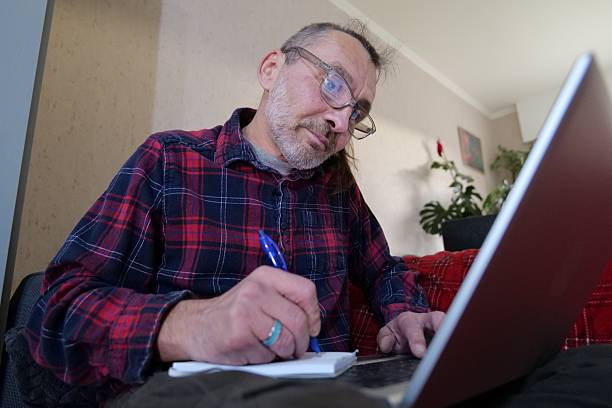 istock Man wearing glasses and a plaid shirt writes in notebook while using laptop 2254085658