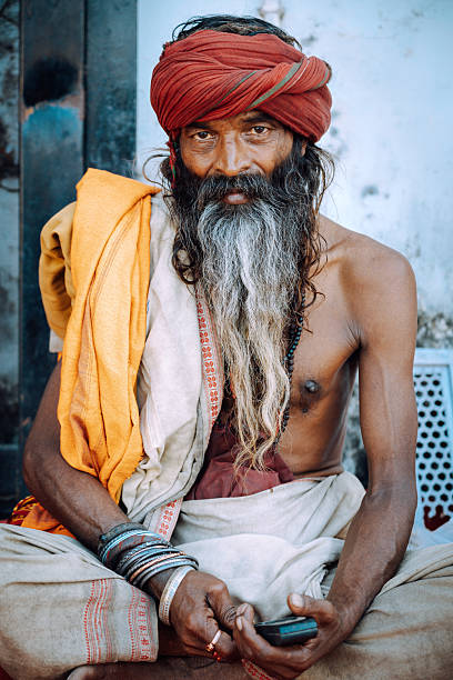 A bearded man with a red turban and flowing gray beard sits with folded hands, draped in orange and cream fabrics. Calm, traditional, and spiritual mood in Pushkar, India.