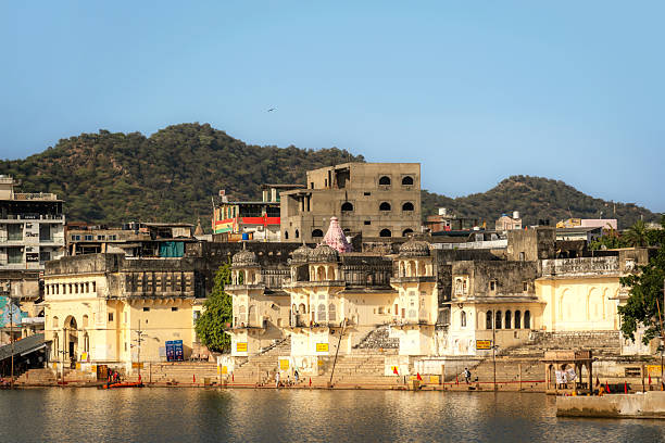 A tranquil view of Pushkar's ancient buildings along the lakefront, with steps descending to the water and distant hills forming a timeless skyline. The scene reflects sacred town life and rich architecture.