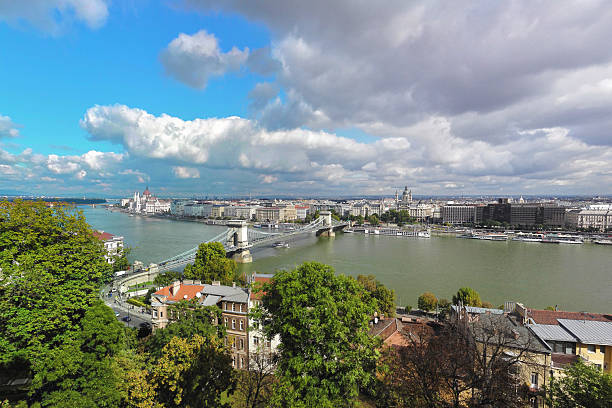 This high-angle scenic view captures the heart of Budapest, showcasing the historic Sz?chenyi Chain Bridge crossing the Danube River towards the magnificent Hungarian Parliament Building. The image highlights the city's unique architectural beauty and vibrant atmosphere under a bright, partly cloudy sky.