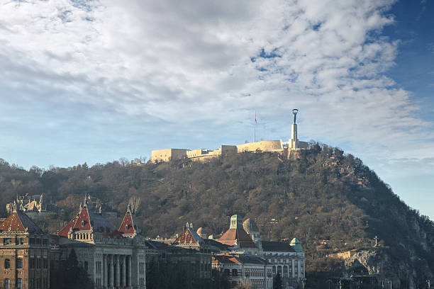 A panoramic view captures the imposing Citadel on Gell?rt Hill in Budapest, Hungary, standing majestically above the city's historic architectural landscape under a partly cloudy sky.