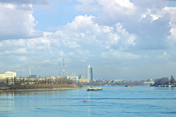 A wide river flows with barges in motion, bordered by a cityscape featuring industrial cranes and a modern skyscraper under a vast, cloudy sky.