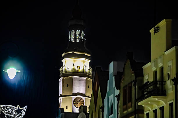 A beautifully lit clock tower stands tall against the dark night sky, surrounded by colorful, old-world buildings