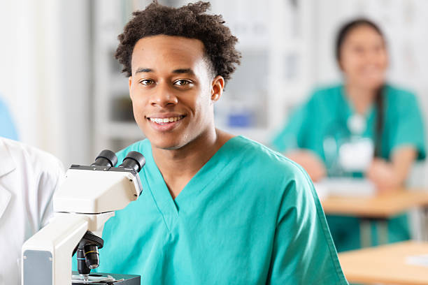A smiling young medical student wearing scrubs looks at the camera, with colleagues in the background. A microscope is in the foreground.