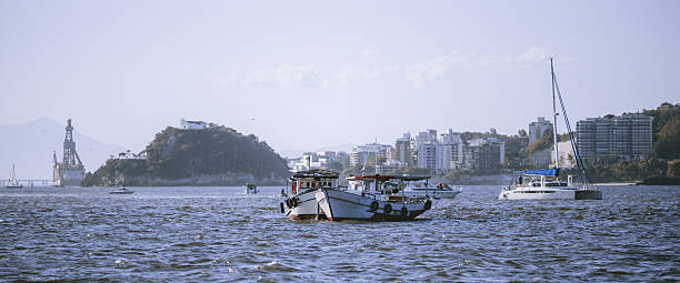 Wide coastal bay with small fishing boat and yacht on rippled water, island hill and city skyline in background, hazy mountains and pale sky, travel and harbor scene