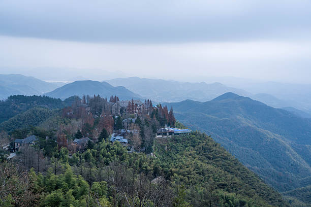 A stunning wide-angle panoramic view overlooking the misty peaks of Moganshan, China. The image shows luxury mountain resorts and villas perched on a ridge, surrounded by dense bamboo forests and rolling hills under a soft, hazy sky. Perfect for travel and environmental themes.