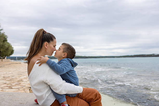 A loving mother sitting by the sea with her young son, sharing a tender moment on a cloudy day.