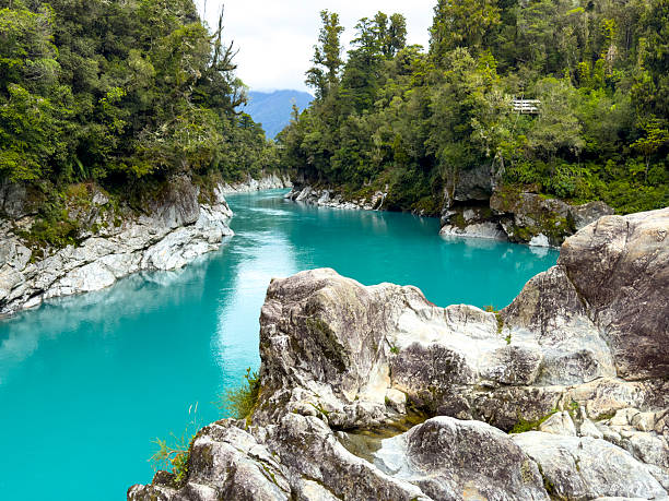 Turquoise water threads through Hokitika Gorge on New Zealand's South Island, framed by lush native forest and rugged rocky banks under a moody cloudy sky, pristine and remote