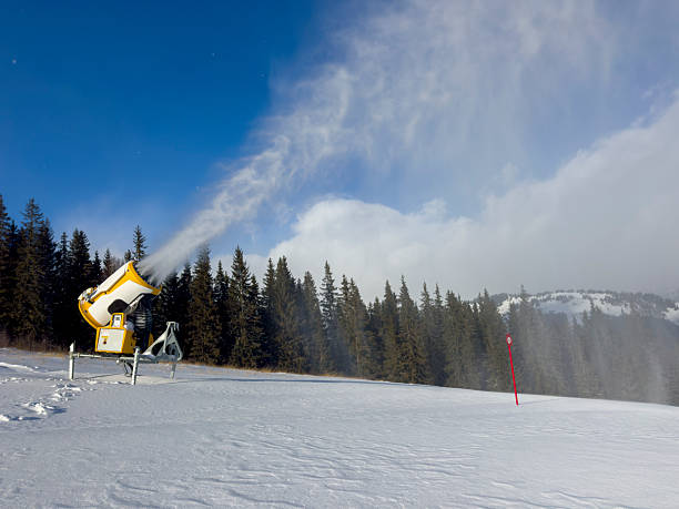 A high-performance snowmaker (fan gun) in action on a groomed ski run, producing fresh artificial snow to ensure optimal conditions for winter sports. The scene is set on a bright, sunny day with a clear blue sky and a backdrop of dense evergreen pine trees. A red slope marker stands on the snow-covered surface, illustrating the professional maintenance and preparation required for the skiing season.