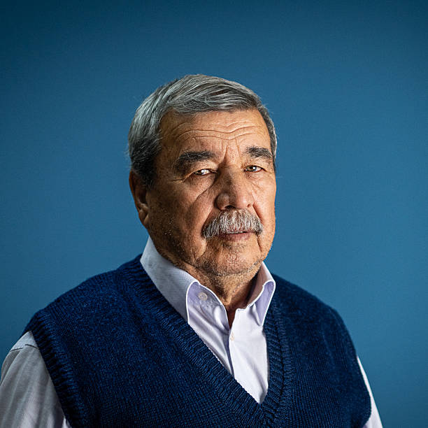 Portrait of a senior Turkish man with gray hair and mustache, wearing a sweater vest and shirt, standing against a blue studio background