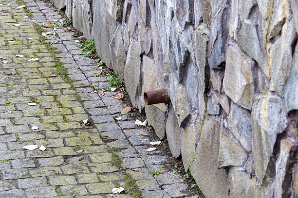 A rusty pipe is sticking out from a stone wall near a cobblestone pathway. Leaves are scattered around the ground.