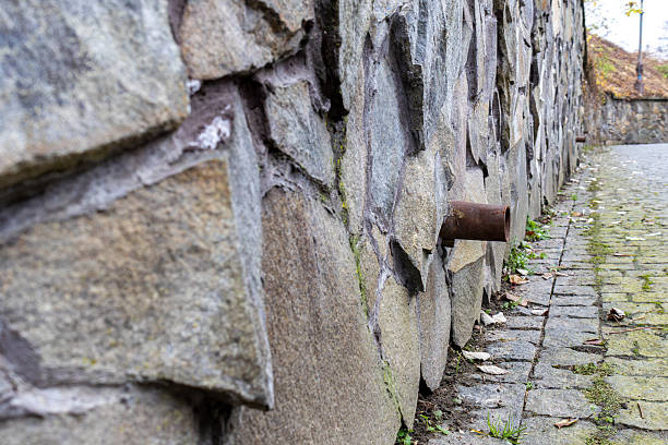 Water pipe sticks out from a stone wall next to a cobblestone path covered with fallen leaves in an urban area.