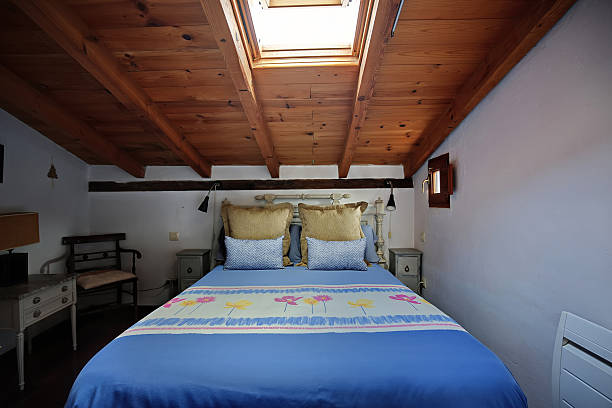 A tranquil attic bedroom featuring a blue bedspread, patterned runners, and soft pillows beneath a warm wooden ceiling and skylight, with vintage furniture creating a cozy, rustic retreat.