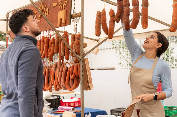 Vendor arranging sausages while customer choosing products at market stall