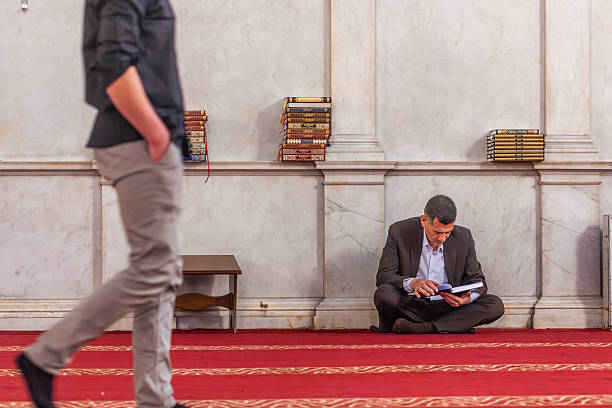 A man sits on the floor of a mosque reading a book while another person walks past him. The setting is simple, with shelves of books and carpeted flooring.