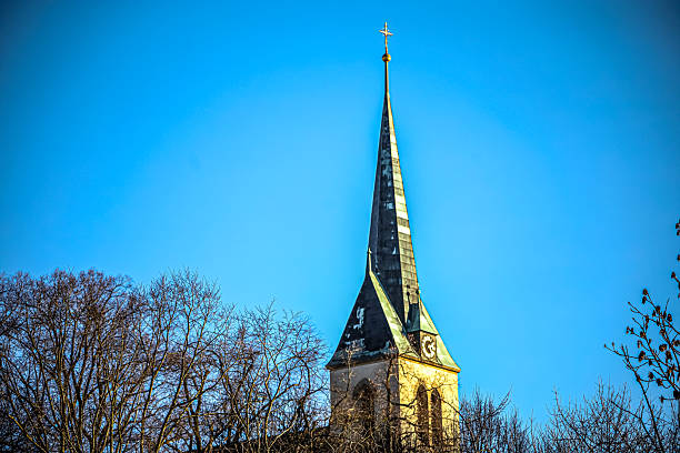 A detailed view of a historic church tower reaching towards the heavens, framed by leafless branches under a bright azure sky