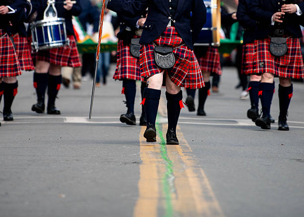 Traditional bagpipers wearing tweed plaid skirts follow the double yellow line in the center of the road as they march in a St. Patrick's Day parade. A green line is painted in the center of the road to celebrate the March 17 holiday.