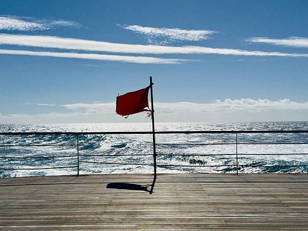 Horizontal view of red flag attached to pole on wooden plank deck. Blue ocean with waves and white foam in background. Clear blue sky with thin clouds. Sunny day, natural lighting.