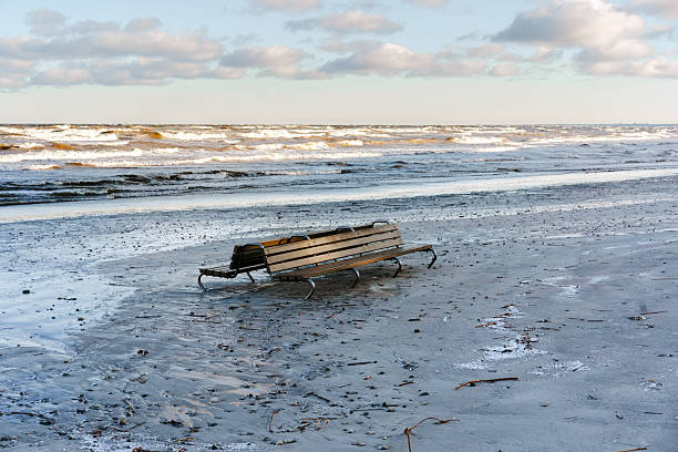 Lonely wooden bench on a wet sandy beach under a cloudy sky. Scenic coastal view with rolling sea waves. Concept of peace, solitude, and nature.