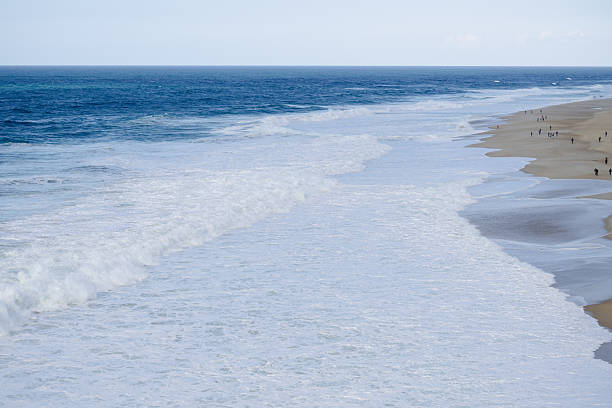 A tranquil coastal scene with foamy waves washing onto a sandy shore. Distant figures enjoy the beach under a clear blue sky, conveying calm, relaxation, and summer vacation vibes.