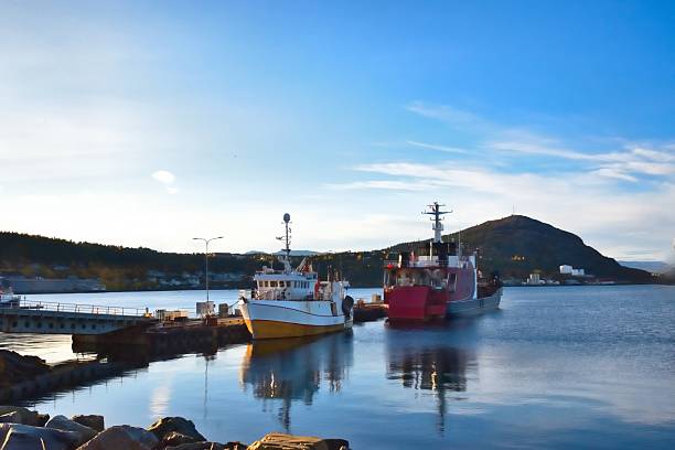 Two boats are tied at a dock in Norway. The water reflects the bright sky and scenery. A mountain is visible in the background, suggesting a remote travel destination.