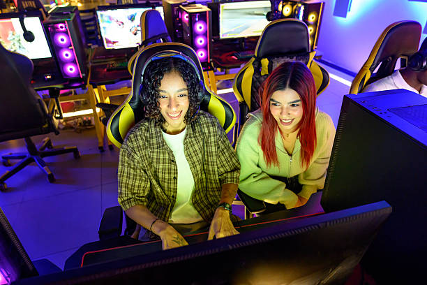 Two young adult female gamers smiling and collaborating while playing a video game at a cyber cafe with glowing neon lights and high-performance computers